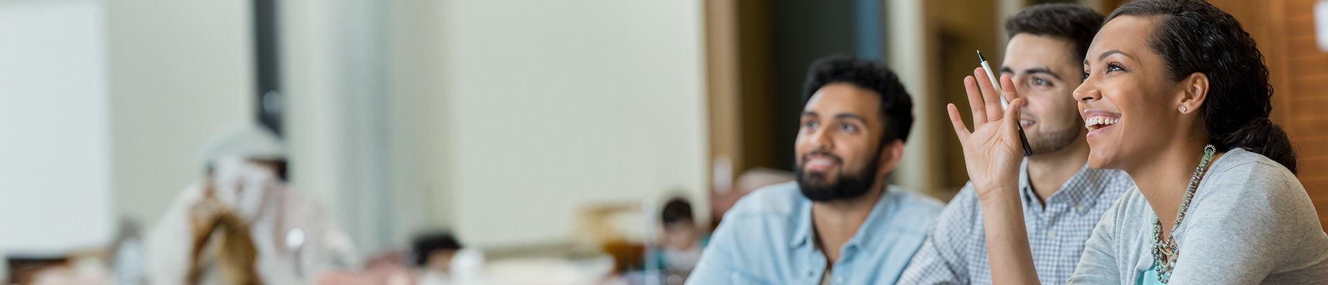 Young woman in a classroom next to two male students, smiling while raising her hand