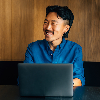 A cheerful businessman in a blue shirt works on his laptop, enjoying a brief pause in a modern office setting with plant decor.