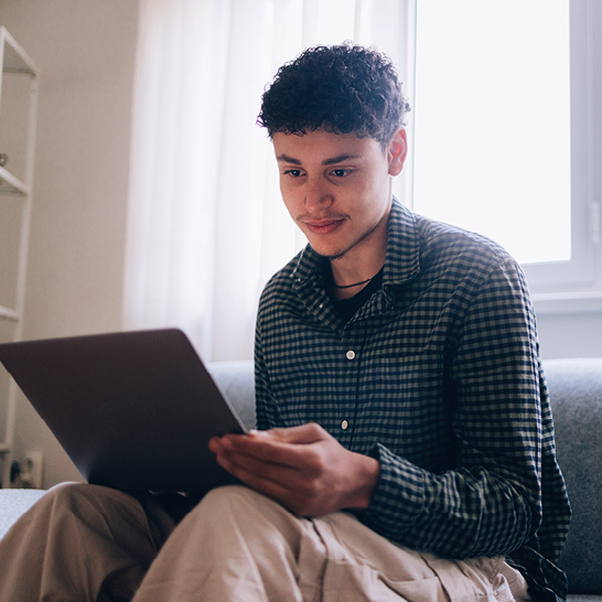 A young man in casual clothing attentively uses his laptop while seated on a sofa.